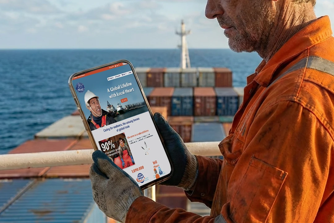 A seafarer stands on the deck of a freight hauler and looks at the mission to seafarers canada website on his tablet